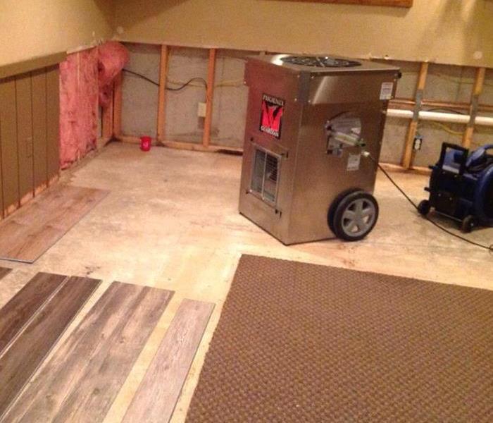 A large air dryer sits in the middle of a basement floor that has had portions of carpeting removed.